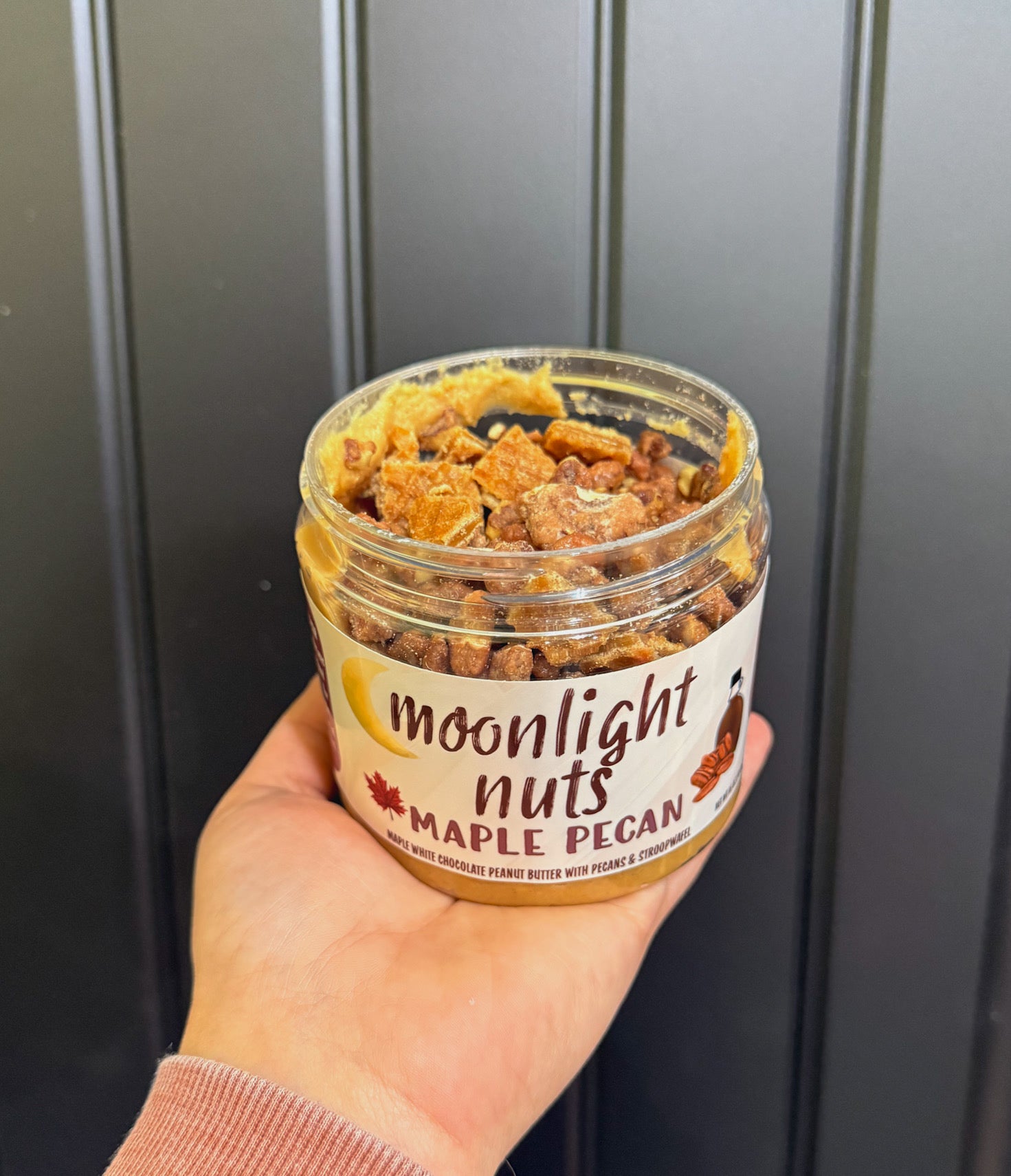 Hand holding a jar of 'Moonlight Nuts Maple Pecan' showing all of the candied pecans and stroopwafel toppings against a dark background