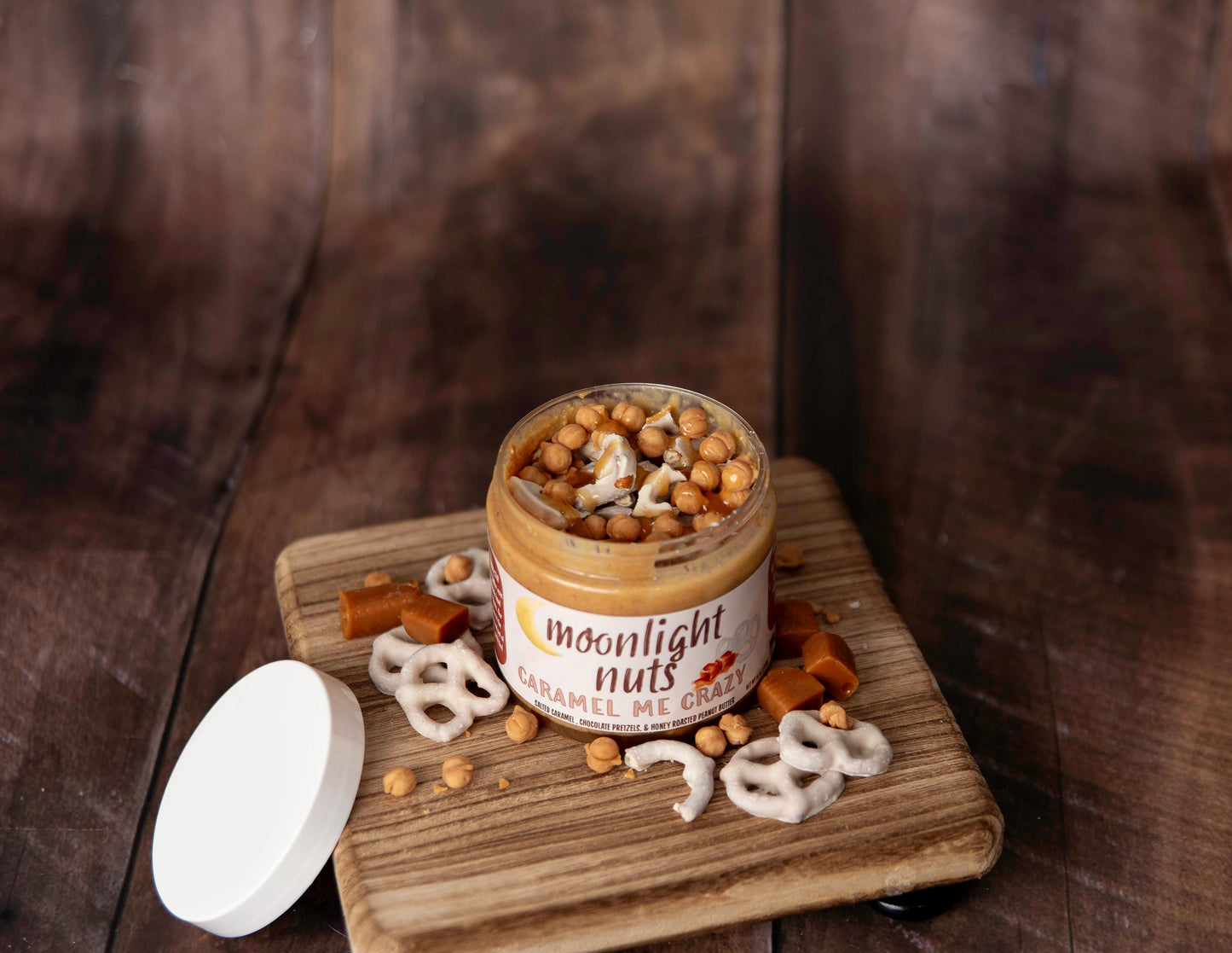 Open jar of moonlight nuts Caramel Me Crazy gourmet peanut butter showing all of the toppings: white chocolate pretzels, caramel bits, salted caramel sauce all on a honey roasted peanut butter base