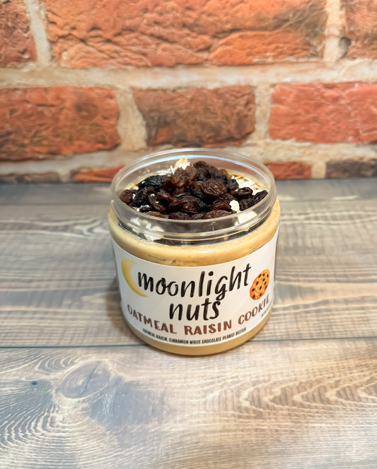 Jar of 'Moonlight Nuts' oatmeal raisin cookie peanut butter spread on a wooden surface with a brick wall background.