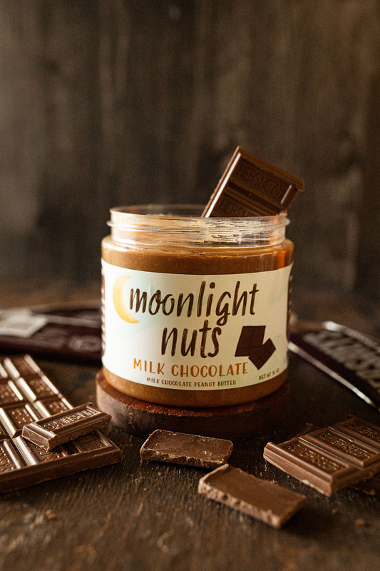A jar of Moonlight Nuts Milk Chocolate peanut butter sits on a dark wooden surface with chocolate bars broken around it. The jar has a white label with brown text, and a chocolate bar is resting inside the jar.